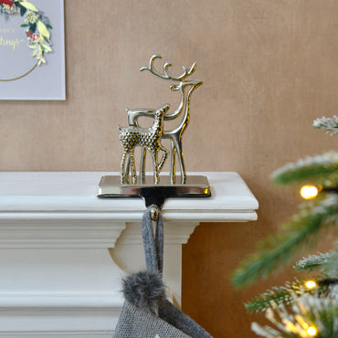 Mr Crimbo double reindeer silver metal Christmas stocking hanger on a mantelpiece with grey Christmas stocking, beside a lit Christmas tree