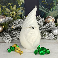 Gonk shaped Christmas storage jar in white with silver detail on a wooden table with green and gold wrapped sweets, snow garland, baubles and fairy lights