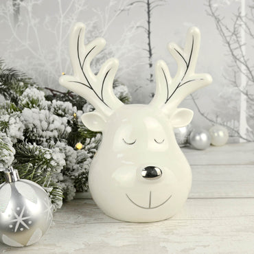 White ceramic reindeer ornament with silver detail, eyes closed on a wooden table with silver Christmas tree baubles, snowy garland and fairy lights