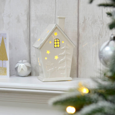 White Christmas house ornament lit up with warm white LED lights on a mantelpiece with silver bauble, christmas card, wood panels and Christmas tree with fairy lights