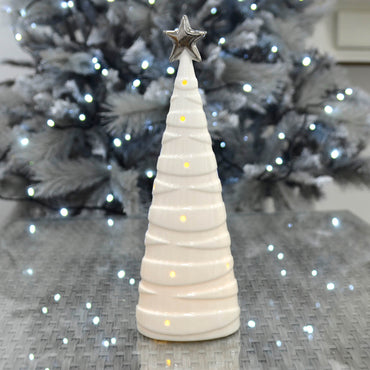 White Christmas tree shape ceramic ornament lit by warm white LEd lights on a grey glass topped table reflecting ice white fairy lights