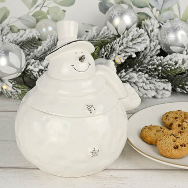 Snowman shape white ceramic Christmas storage jar with silver detail on a wooden table with chocolate chip cookies on a plate, silver baubles, pine garland and LED lights