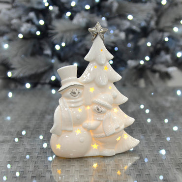 White snowmen and Christmas tree ceramic ornament with silver detail, sitting on a glass topped table reflecting ice white lights from a silver Christmas tree