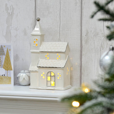 White ceramic town house Christmas ornament with silver detail lit by warm white LED lights, sitting on a mantelpiece with silver bauble and Christmas tree