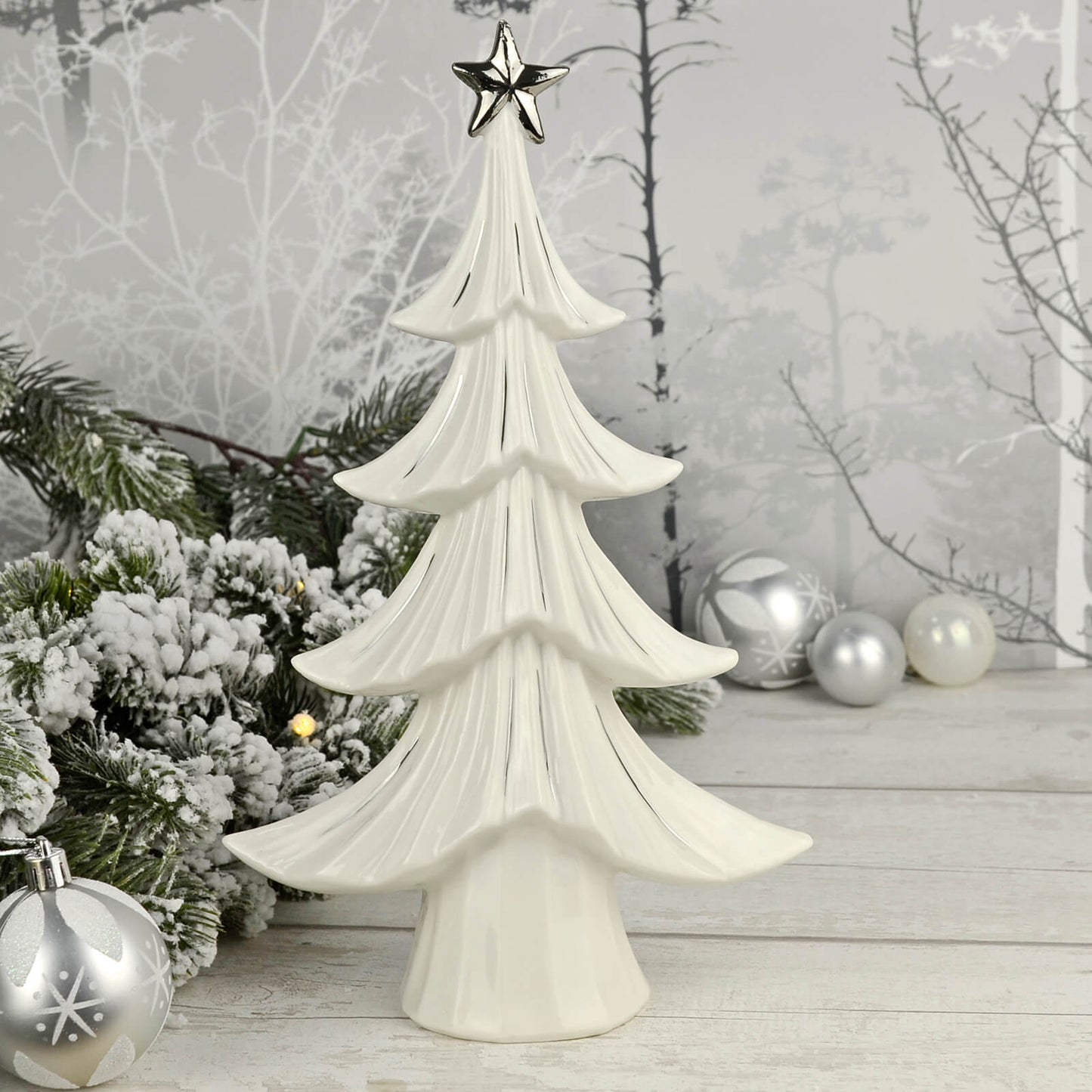 Christmas tree shaped white ceramic ornament with silver star and details on a wooden table with pine garland, silver baubles and LED lights