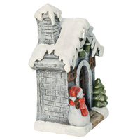 Side view of Christmas ornament with snowman and tree at the door of a snow covered, grey brick house with Christmas wreath
