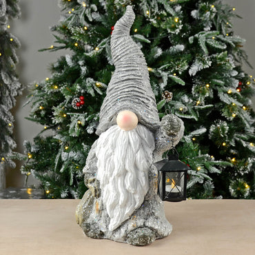 Grey and white sparkling Christmas gonk holding a black metal tealight lantern standing on a table with lit Christmas tree