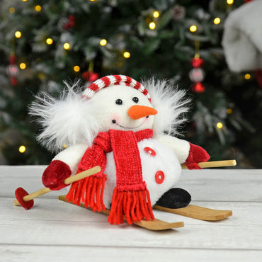 Snowman on skis Christmas decoration with plush red scarf, ear muffs, black felt boots, wooden skis and ski poles, sitting on a wooden table with lit Christmas tree