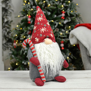 Christmas gonk with grey and red striped candy cane, red knitted hat with snowflakes, and a fluffy white beard, sitting on a wooden table with decorated Christmas tree