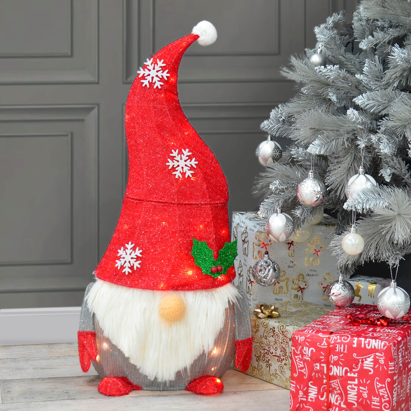 Light up Christmas gnome with snowflake red glitter hat and holly leaves sitting beside a silver Christmas tree and presents