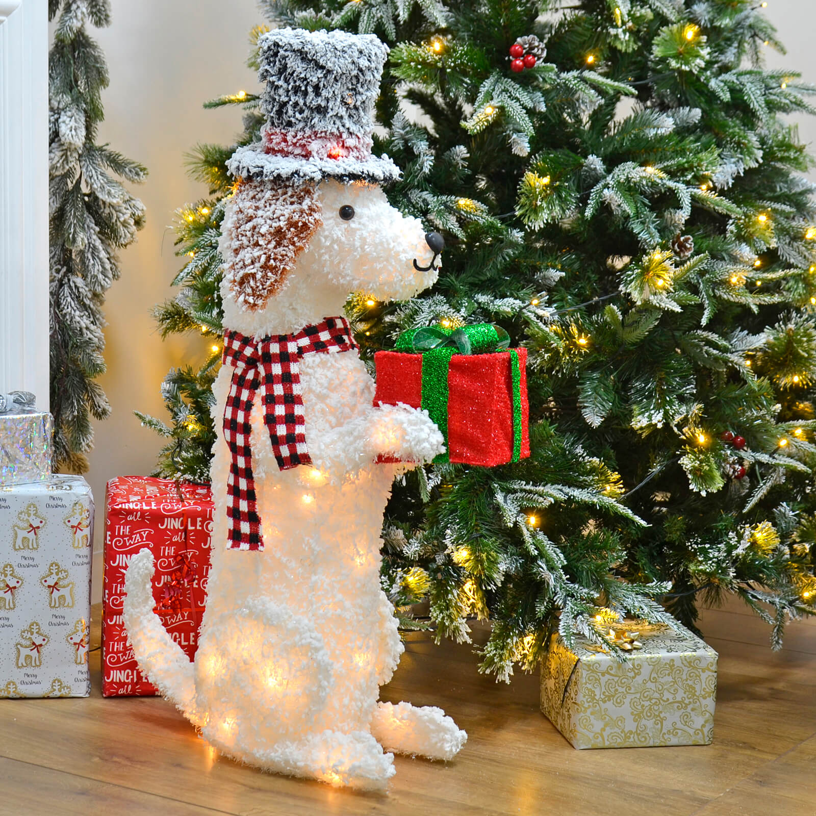 Large white snow dog Christmas ornament with scarf and top hat, holding a red and green Christmas present beside a lit Christmas tree