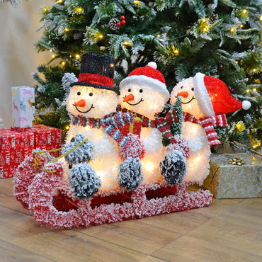 Three snowmen on a red sledge light up Christmas decoration sitting in front of a green pine Christmas tree and presents