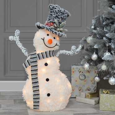 Happy snowman light upChristmas decoration with grey and white scarf, top hat with pine foliage, sitting beside a silver Christmas tree with presents