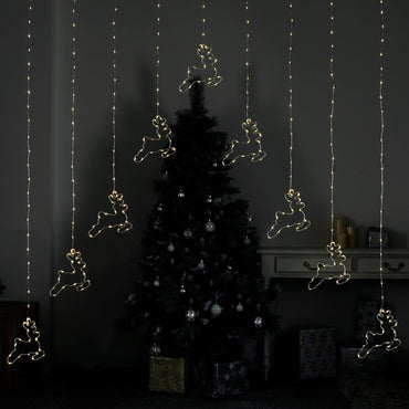 Christmas curtain light with 9 reindeer shapes lit by warm white LED lights, hanging in front of a Christmas tree and presents