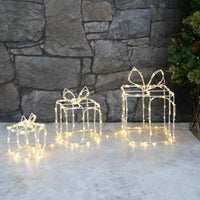 Set of 3 light up Christmas presents outdoor decoration with warm white LEd lights on white wire frame on a patio with Christmas tree