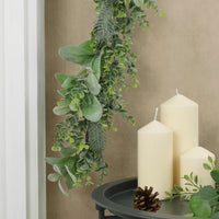 mr crimbo mixed green foliage christmas garland hanging over fireplace mantel beside grey metal table featuring pillar candles and pine cone