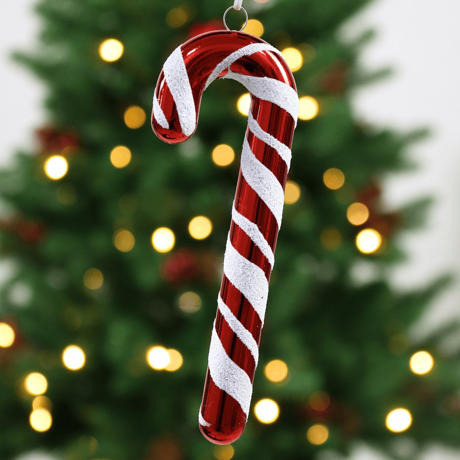 red glossy candy cane decoration with white glitter stripes hanging in front of christmas tree with warm white led lights